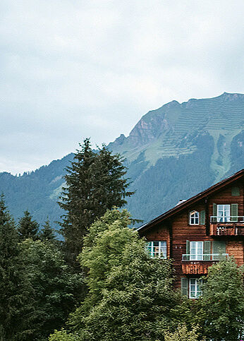 View of an old wooden house partially obscured by green trees in the Swiss Alps on a cloudy summer day.